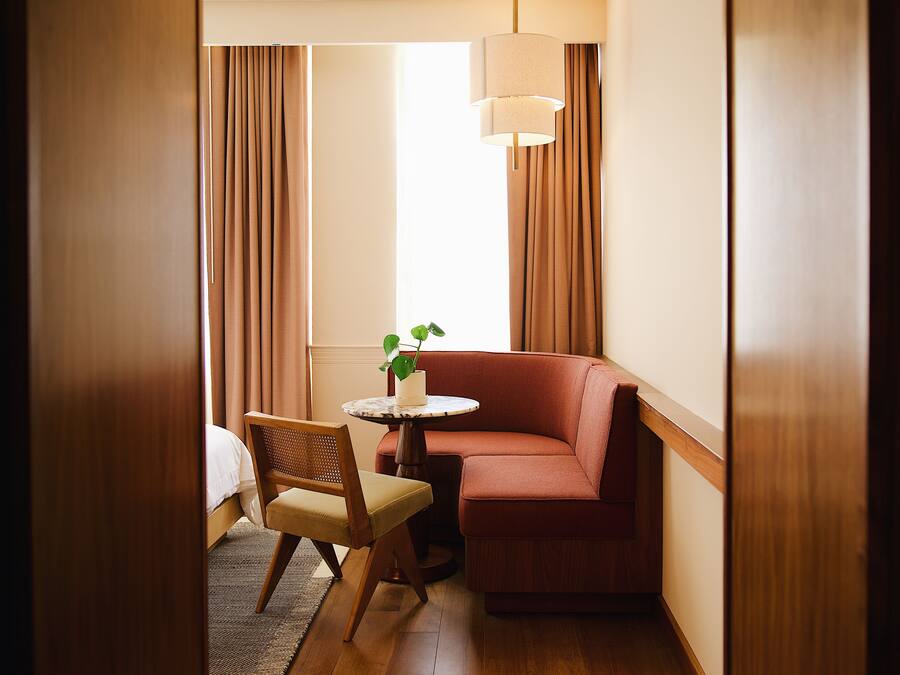 A cozy hotel room with a wooden floor, a red sofa, a small round table with a plant, and a chair.