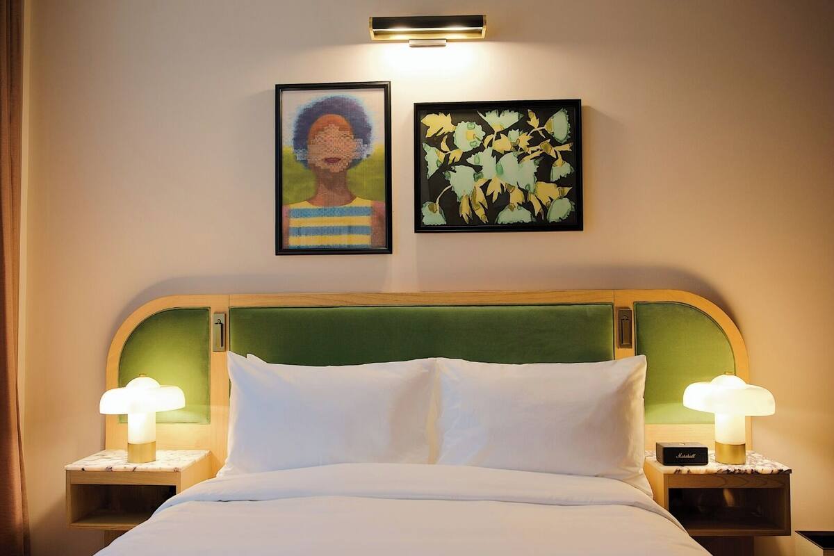 A hotel room with a bed, two nightstands, two lamps, a green upholstered headboard, and two framed artworks on the wall.
