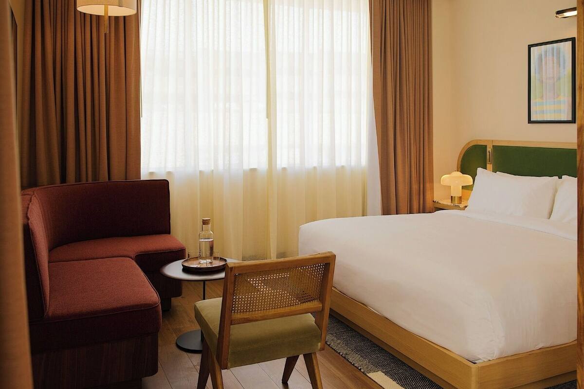 A hotel room with a bed, a red armchair, a wooden chair, a small table with a bottle and glasses, and a nightstand with a lamp.