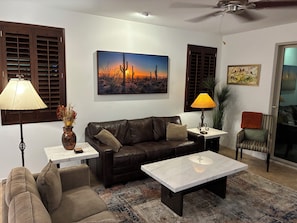 Un salon avec un canapé en cuir marron, une table basse blanche, un tapis à motifs, un tableau représentant un paysage désertique et un ventilateur de plafond.