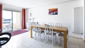 A dining area with a wooden table and white chairs, a red and white abstract painting, and a view of the outside balcony. - La Belle Escapade - Climatisée, aperçu mer (Port-Vendres)