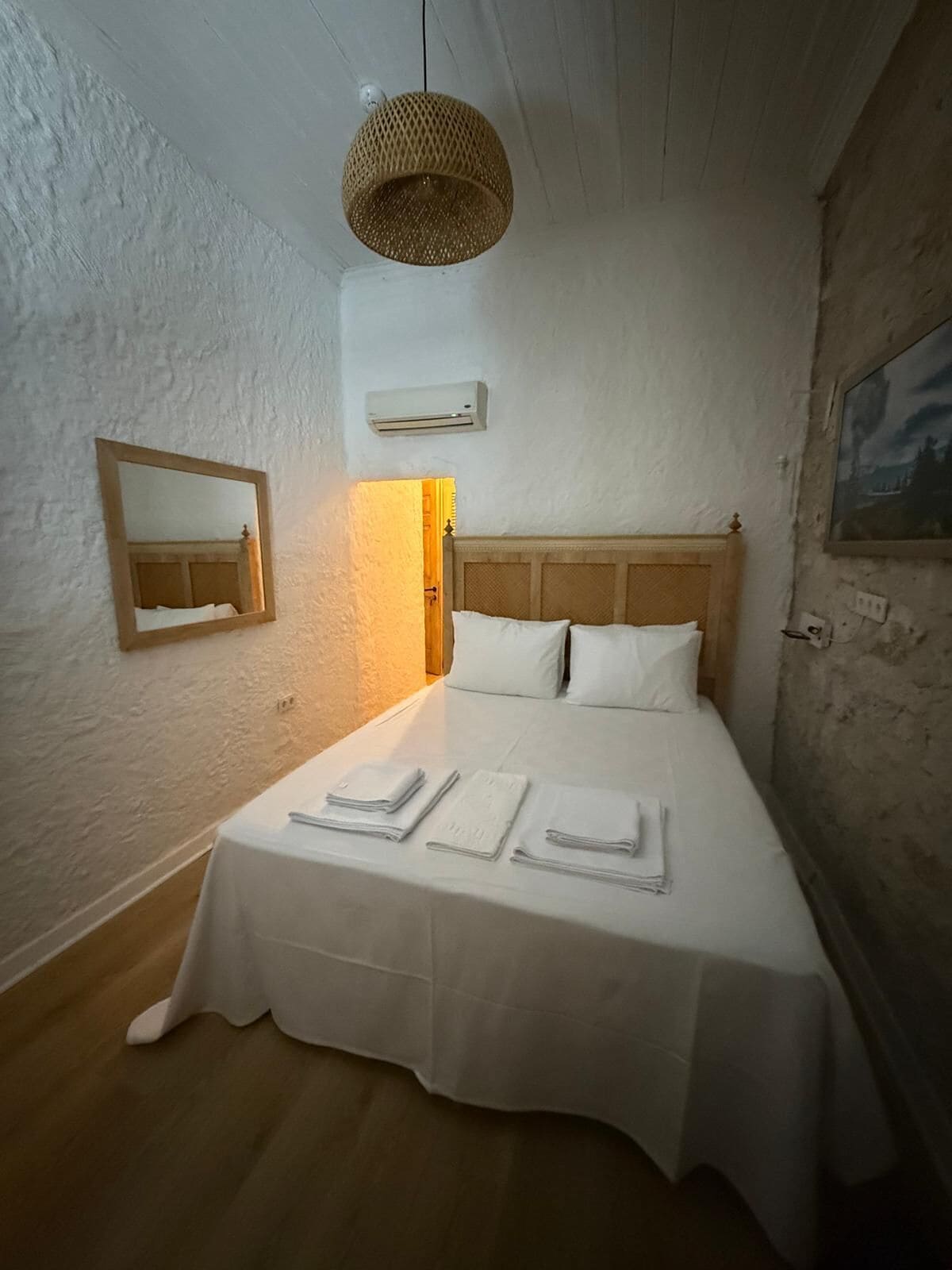 Un lit bien fait, avec du linge de lit et des serviettes blanches, une tête de lit en bois, un luminaire suspendu en fibres tressées, un climatiseur fixé au mur et un miroir encadré.