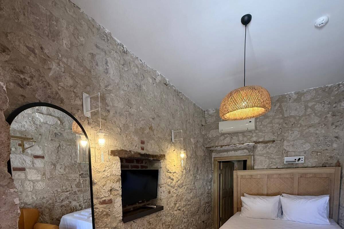 Un dormitorio con pared de piedra, cama con ropa de cama blanca, cabecero de madera, televisor montado en la pared y lámpara de techo tejida.