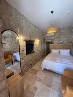 Un dormitorio con pared de piedra, cama con ropa de cama blanca, cabecero de madera, televisor montado en la pared y lámpara de techo tejida.