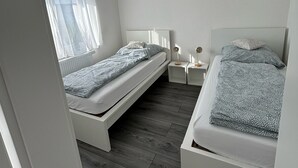 A bedroom with two beds, a window with sheer curtains, a ceiling fan, and a bedside table with a lamp. - Vidi Apartmens: By the stream (Bad Überkingen)