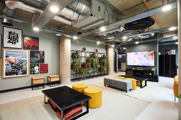 A modern lounge area with a black table, yellow ottomans, a large TV, and artwork on the walls. - Student Only Vita St. Albans (Leeds)