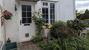 Une maison blanche avec une porte blanche, deux fenêtres et un petit porche agrémenté de plantes en pot et d’un bac vert.