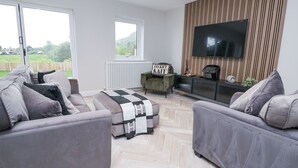 A modern living room with two grey sofas, a coffee table, a TV mounted on a wooden panel wall, and a view of greenery through large windows. - Ty Blodau (Prestatyn)