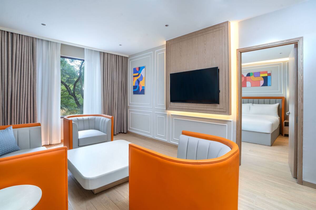 A modern hotel room with a large TV, a sofa, a chair, a bed, and a window with curtains.