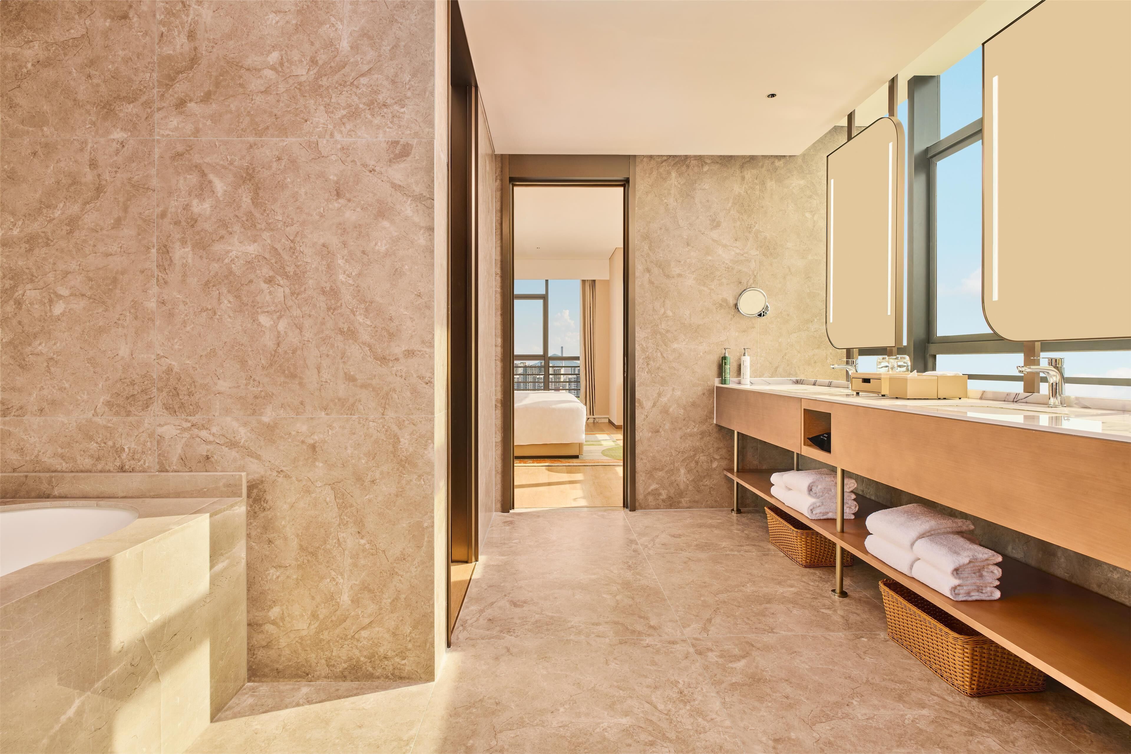 Une salle de bain moderne avec un sol en marbre, un grand miroir, une baignoire et une vue sur une chambre à coucher, visible à travers une porte ouverte.