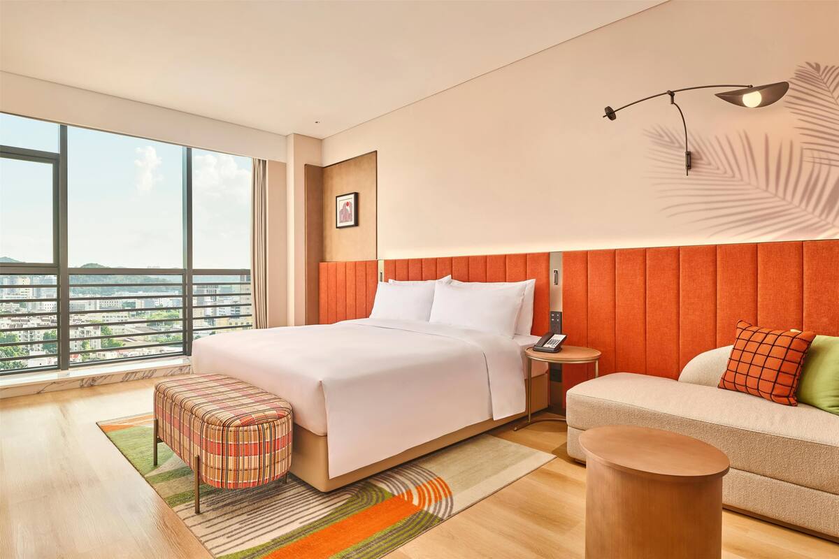 Een moderne hotelkamer met een groot bed, een slaapbank, een klein rond tafeltje en uitzicht op de stad door grote ramen.