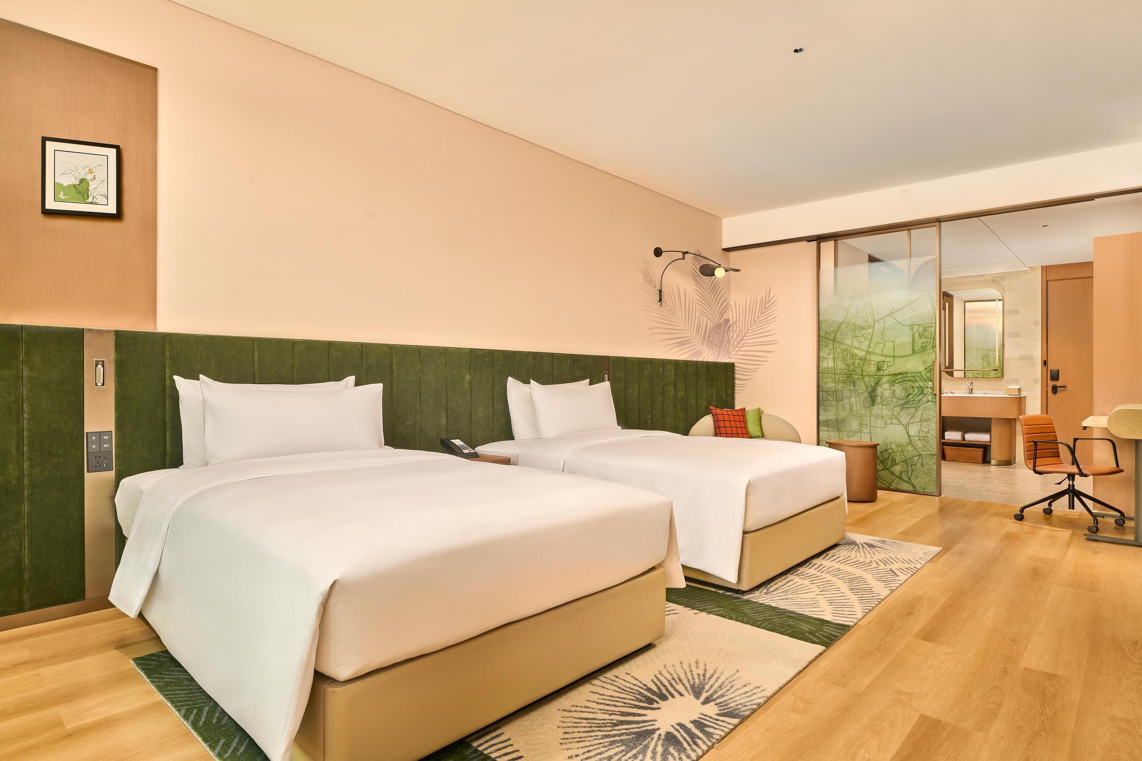Ein Hotelzimmer mit zwei Betten, einem Schreibtisch und einem durch eine Glastür einsehbaren Badezimmer.