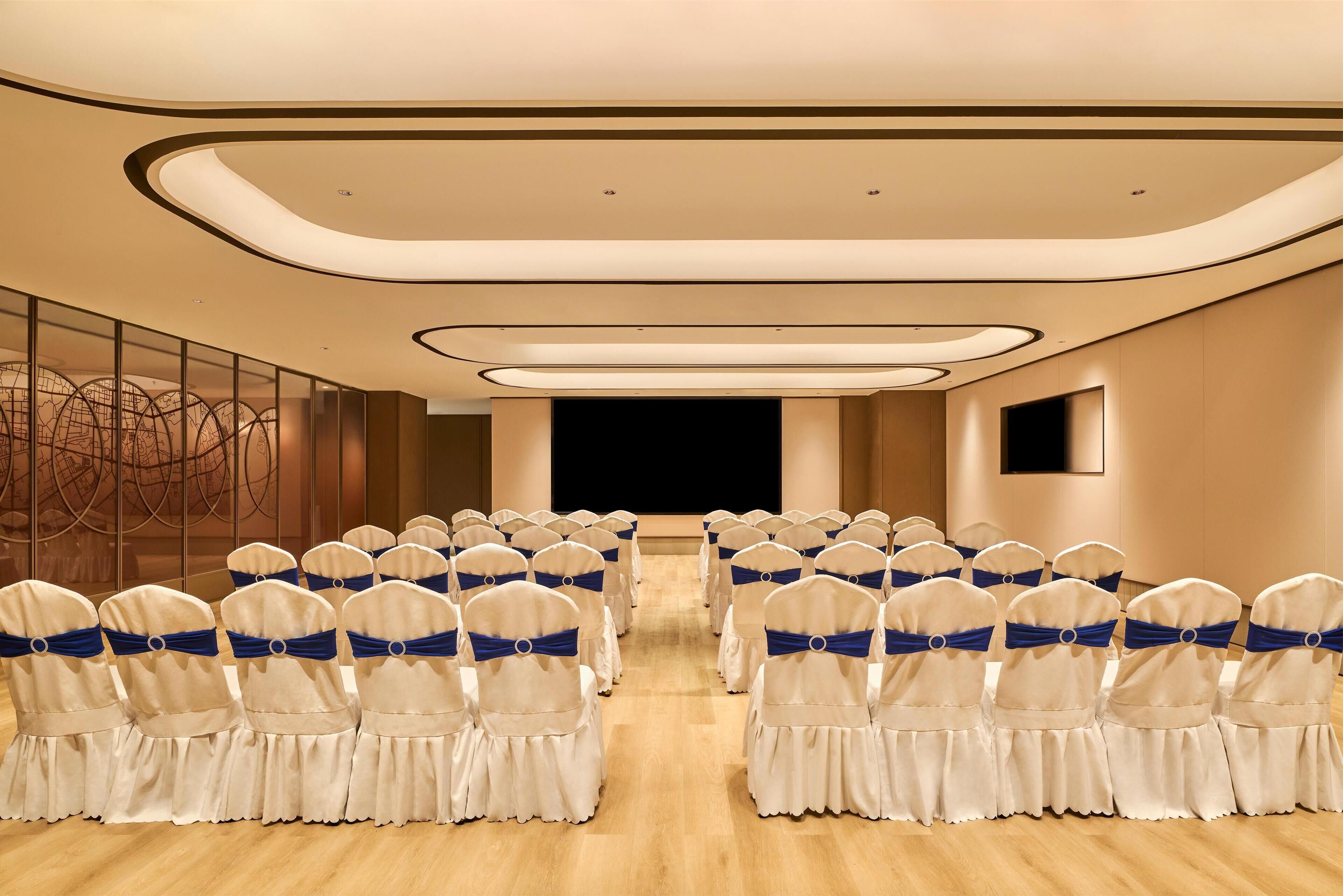 A conference room with rows of white chairs with blue sashes, a stage, and a large screen.