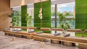A modern lounge area with a long bench, small round tables, and potted plants. - Hilton Garden Inn Zhongshan South (Zhongshan)