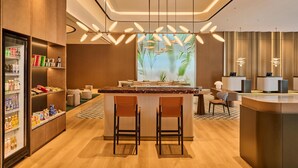A modern hotel lobby with a central bar area, seating, a refrigerator stocked with beverages, and a large window with a view of palm trees. - Hilton Garden Inn Zhongshan South (Zhongshan)