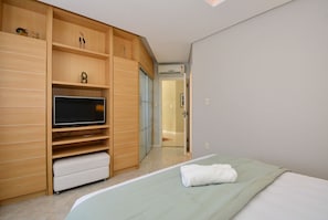 Une chambre à coucher avec un lit, un téléviseur posé sur une étagère en bois, un banc blanc et une porte donnant sur une autre pièce.