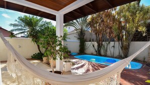 Eine Hängematte unter einer Holzpergola, ein Schwimmbecken und verschiedene Kübelpflanzen.