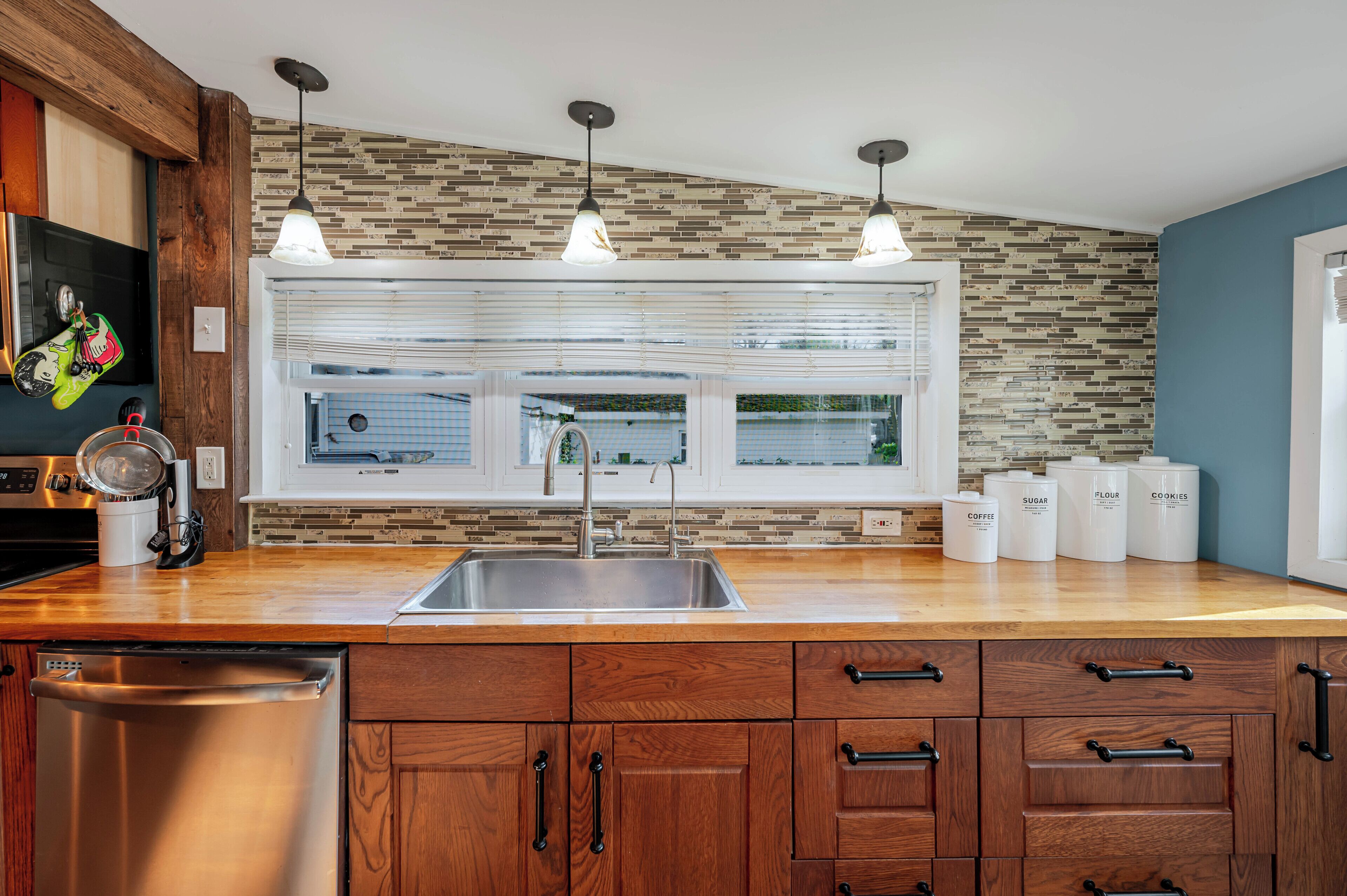 A modern kitchen with a wooden countertop, stainless steel sink, and a window with blinds. There are three hanging pendant lights above the sink.