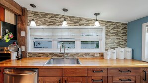 A modern kitchen with a wooden countertop, stainless steel sink, and a window with blinds. There are three hanging pendant lights above the sink.