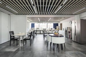 Un espace repas moderne avec plusieurs tables et chaises, un coin cuisine équipé d’un réfrigérateur et une vue par la fenêtre.