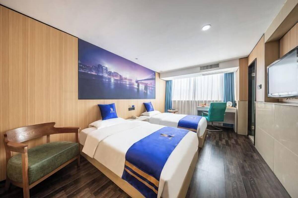 A hotel room with two beds, a desk, a chair, a TV, and a large window with curtains.
