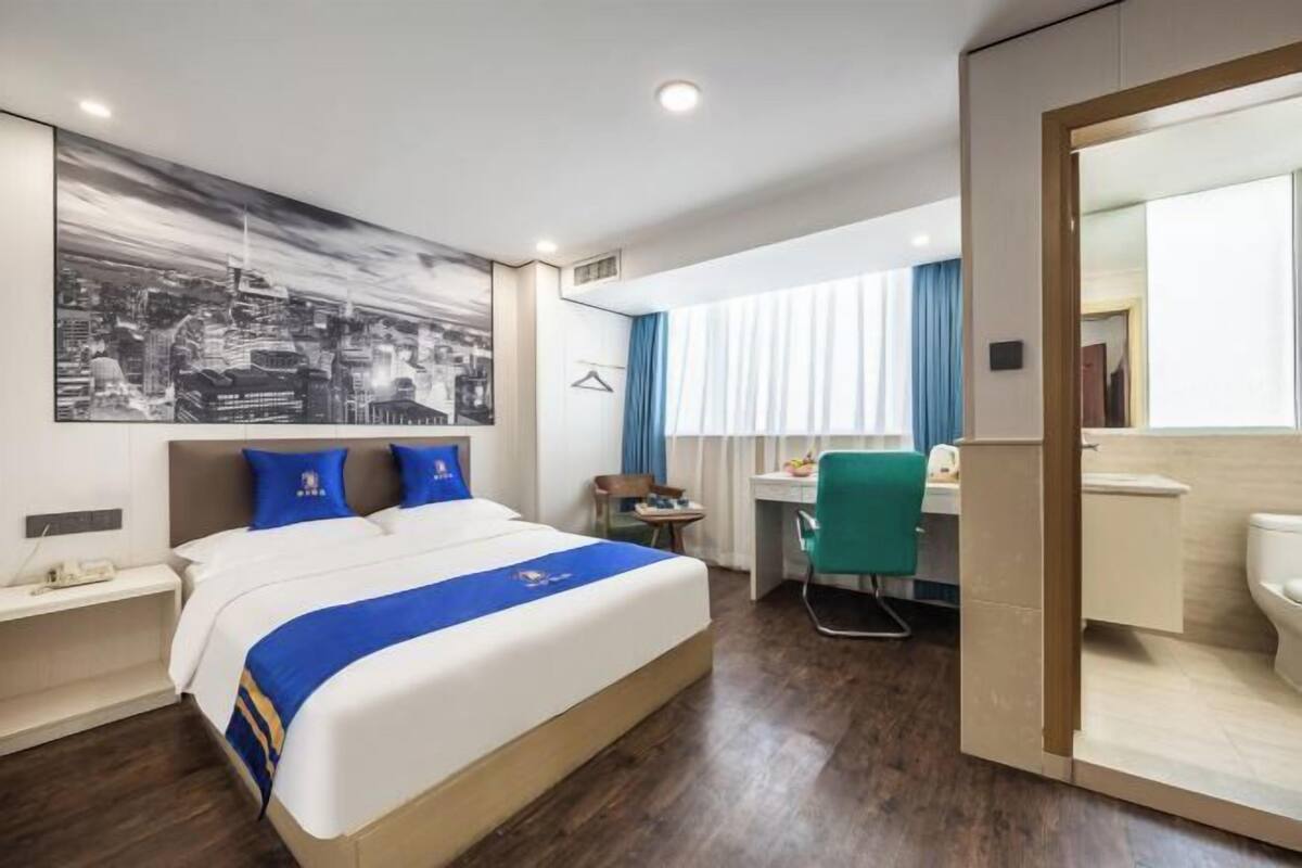 A hotel room with a bed, a desk, a chair, a bathroom, and a cityscape artwork on the wall.