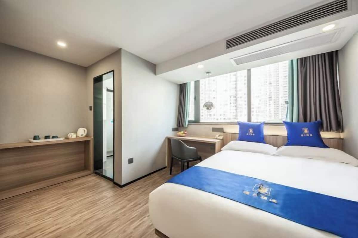 A hotel room with a bed, desk, chair, and a view of a cityscape through the window.