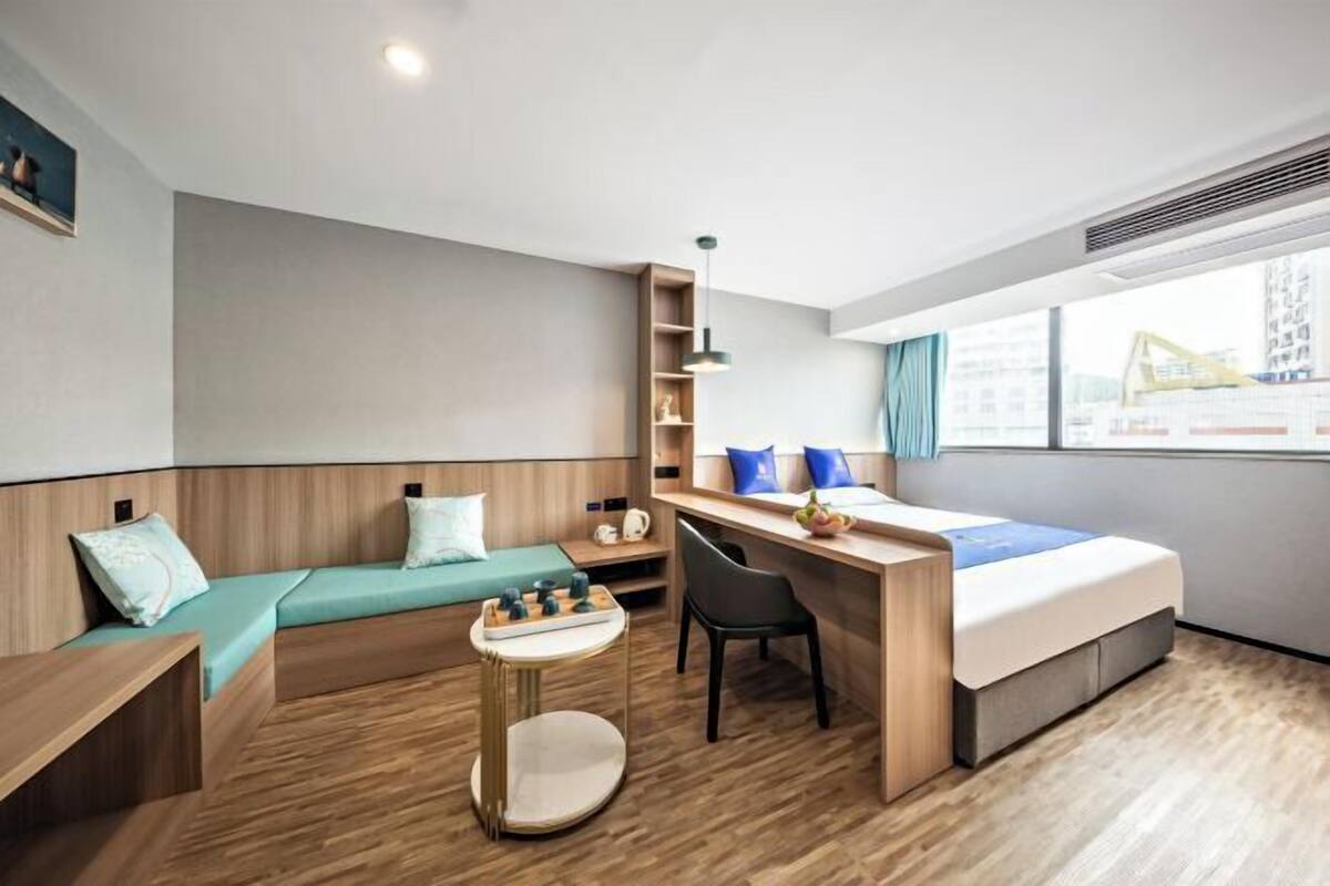 A modern hotel room with a wooden floor, a bed with a blue bedspread, a desk with a chair, a small round table, and a shelf with decorative items.