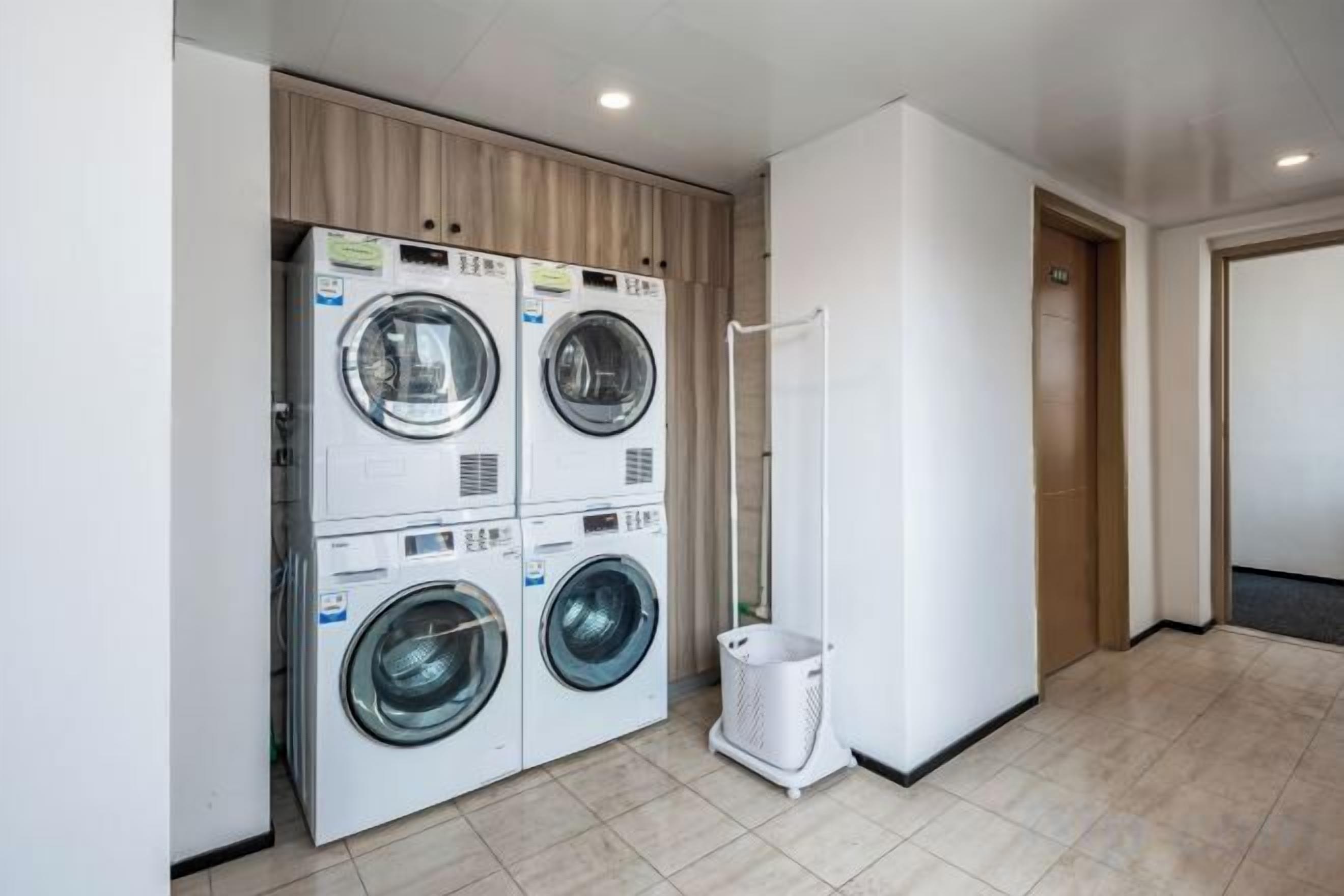 Un lavadero con lavadoras y secadoras apiladas, un bote de basura blanco y una puerta que da a otra habitación.