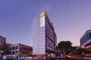 Un hotel de varios pisos con un logotipo destacado en la fachada, situado en un entorno urbano con edificios circundantes.