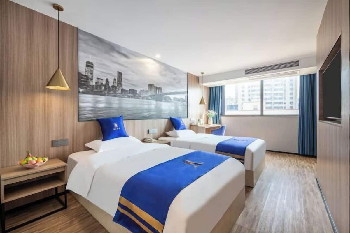A hotel room with two beds, a cityscape painting, a fruit bowl, and a large window.