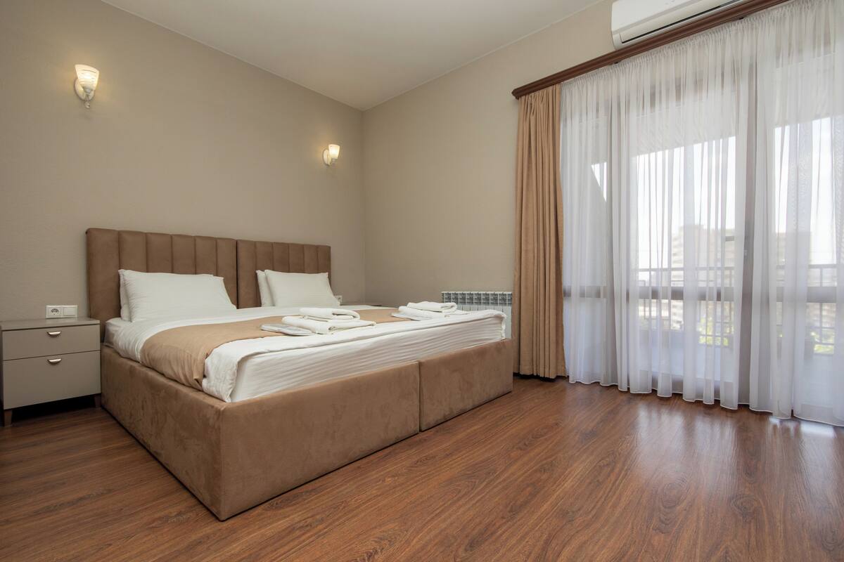 Una habitación de hotel con una cama grande, piso de madera, una mesita de noche y una ventana con cortinas ligeras.