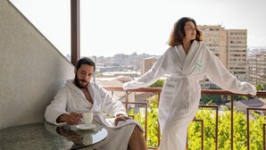 A balcony with a city view, a round table with a cup and saucer, and two individuals in white robes.