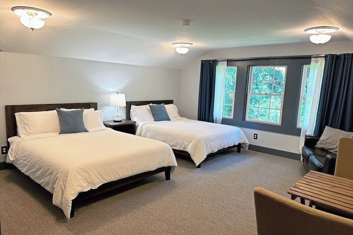 A hotel room with two beds, a desk, and a chair. There are windows with curtains, a ceiling fan, and a lamp on the nightstand.