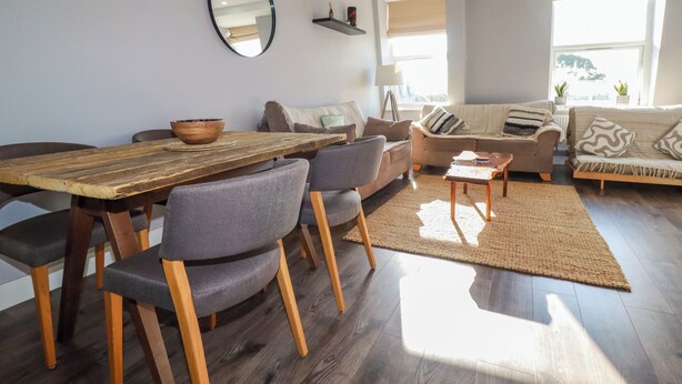 A modern living room with a wooden dining table, grey upholstered chairs, a sofa, and a coffee table. - Priory (St Austell)