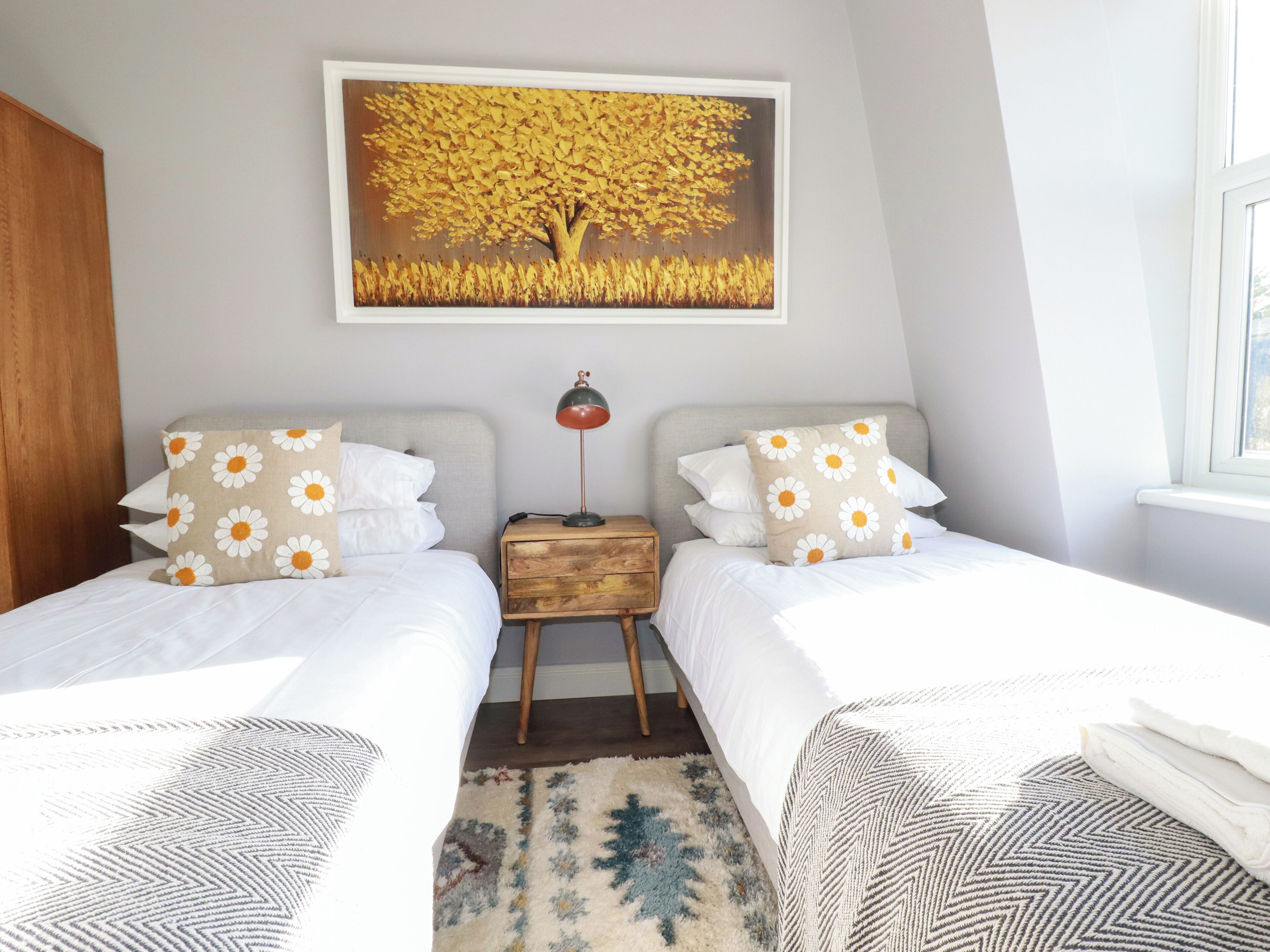 Une chambre avec deux lits, une table de chevet, une lampe, un tableau représentant un arbre et une fenêtre.