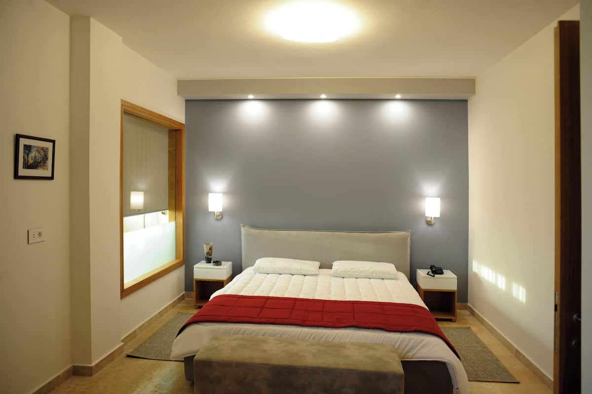 Une chambre d’hôtel avec un grand lit, deux tables de chevet, une petite table d’appoint, un plaid rouge et une fenêtre avec des stores.