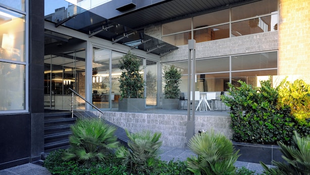 A modern building with large glass windows, a staircase, and outdoor seating area with plants.