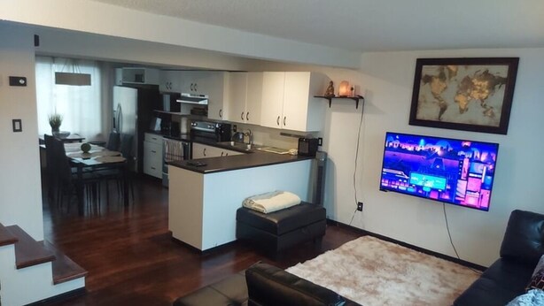 Una sala de estar moderna con un sofá de cuero negro, un televisor de pantalla plana montado en la pared y una cocina al fondo.