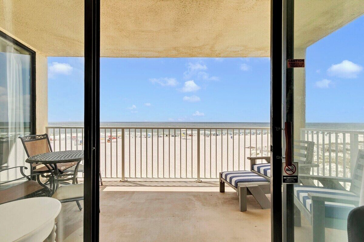 A balcony with a view of the beach, a table, chairs, and a bench.