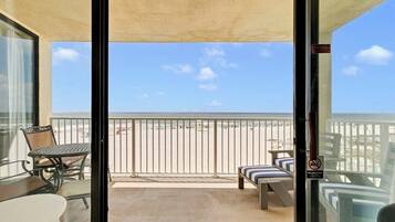 A balcony with a view of the beach, a table, chairs, and a bench.