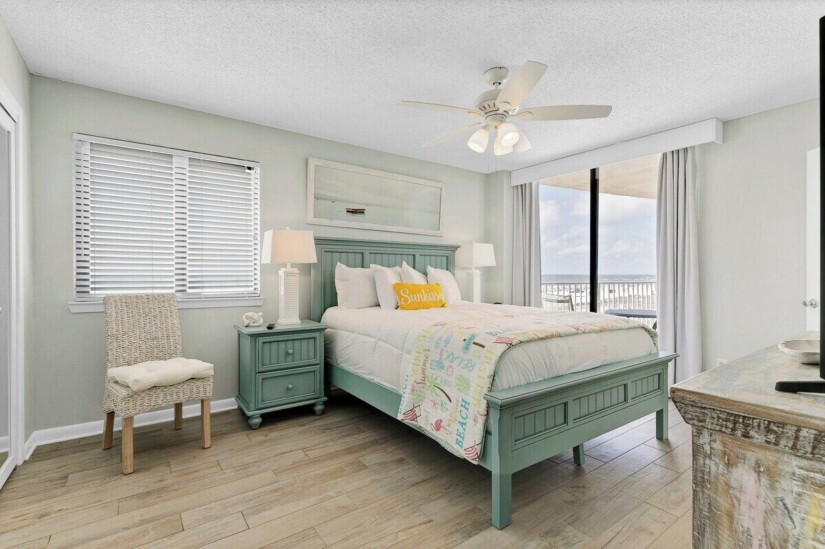 A bedroom with a bed, a chair, a nightstand, a ceiling fan, and a view of the outdoors.
