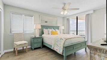 A bedroom with a bed, a chair, a nightstand, a ceiling fan, and a view of the outdoors.