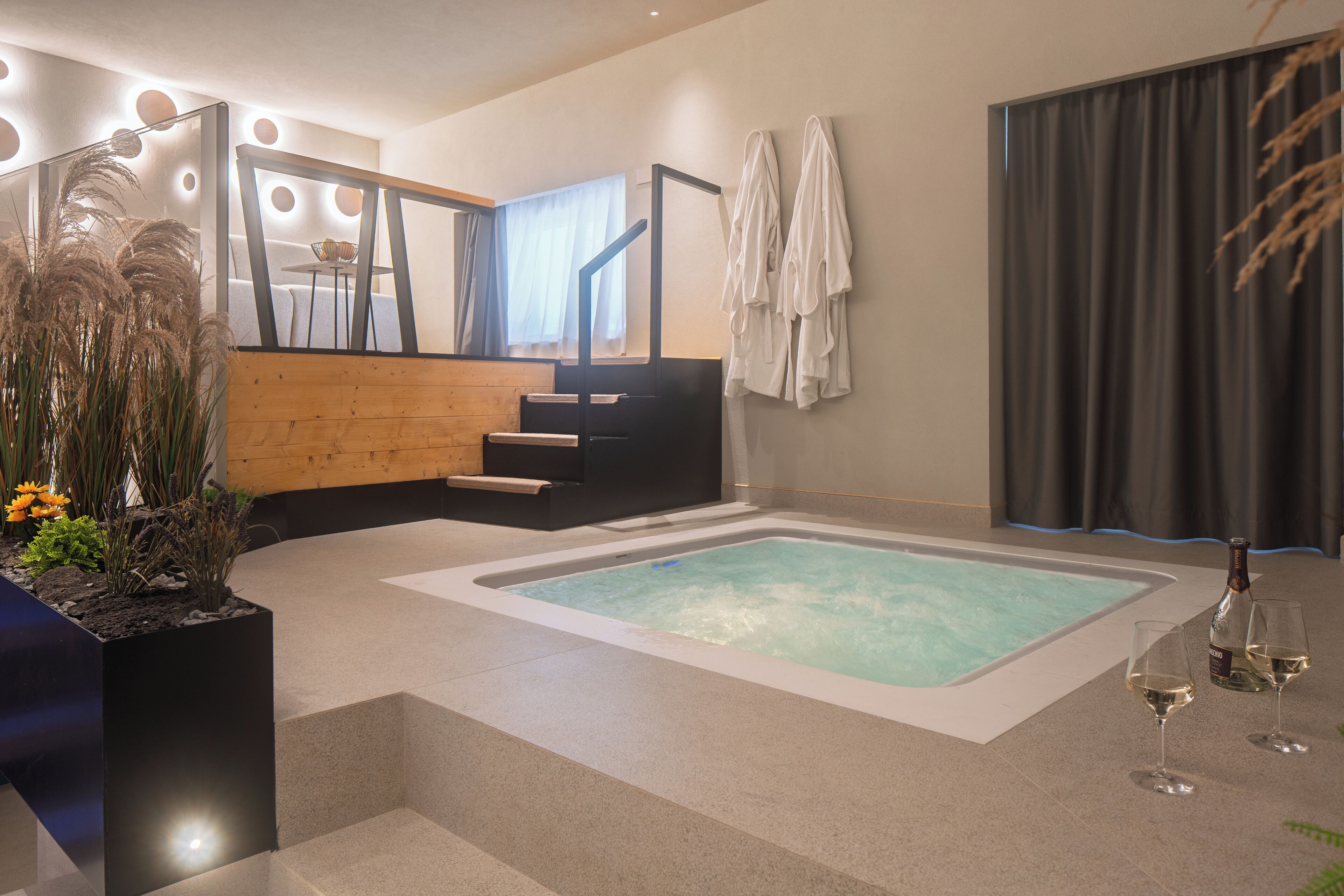 A modern bathroom with a large bathtub, a wooden bench, a mirror with lights, and a bottle of champagne.