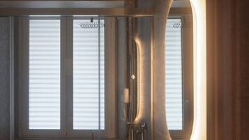 A modern bathroom with a glass-enclosed shower, a round mirror, and a large window with blinds.