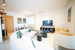 Una sala de estar moderna con un sofá blanco, una mesa de centro negra y un televisor sobre un soporte negro. Se ve una puerta de madera y un comedor al fondo.
