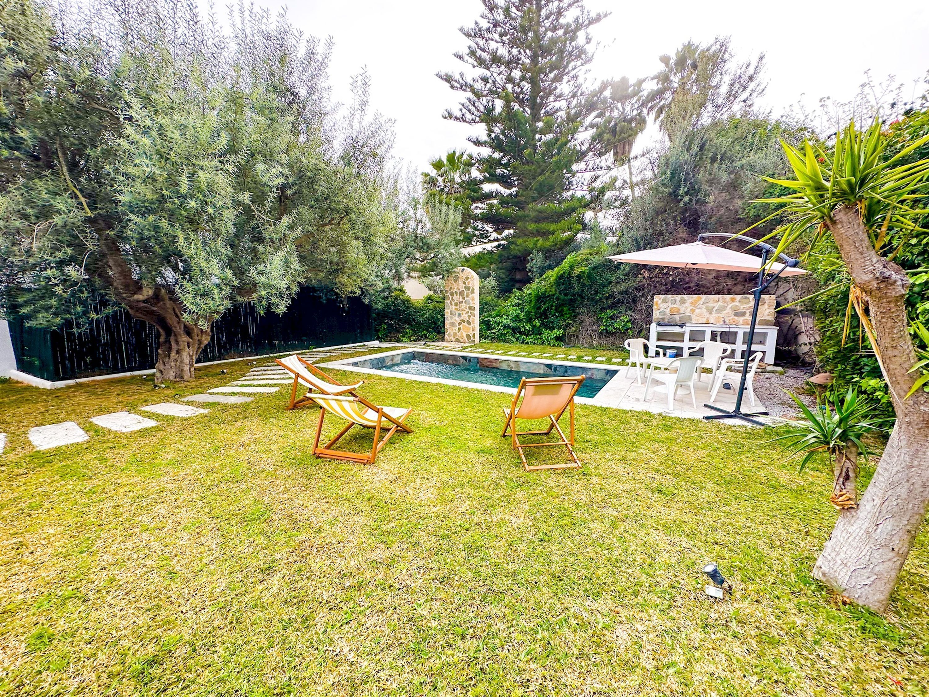 Un patio trasero con piscina, dos sillones de descanso, una zona de barbacoa de piedra y abundante vegetación.