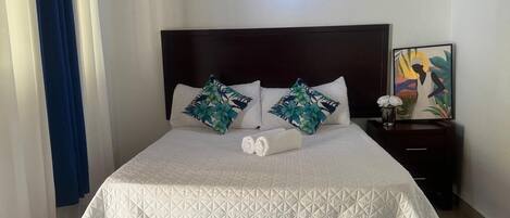 Una cama bien hecha con una colcha blanca acolchada, dos almohadas con estampado de hojas tropicales y una alfombra mullida blanca al pie de la cama.