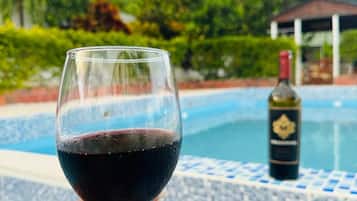 Une main tenant un verre à vin rempli de vin rouge, avec une bouteille en arrière-plan près d’une piscine.