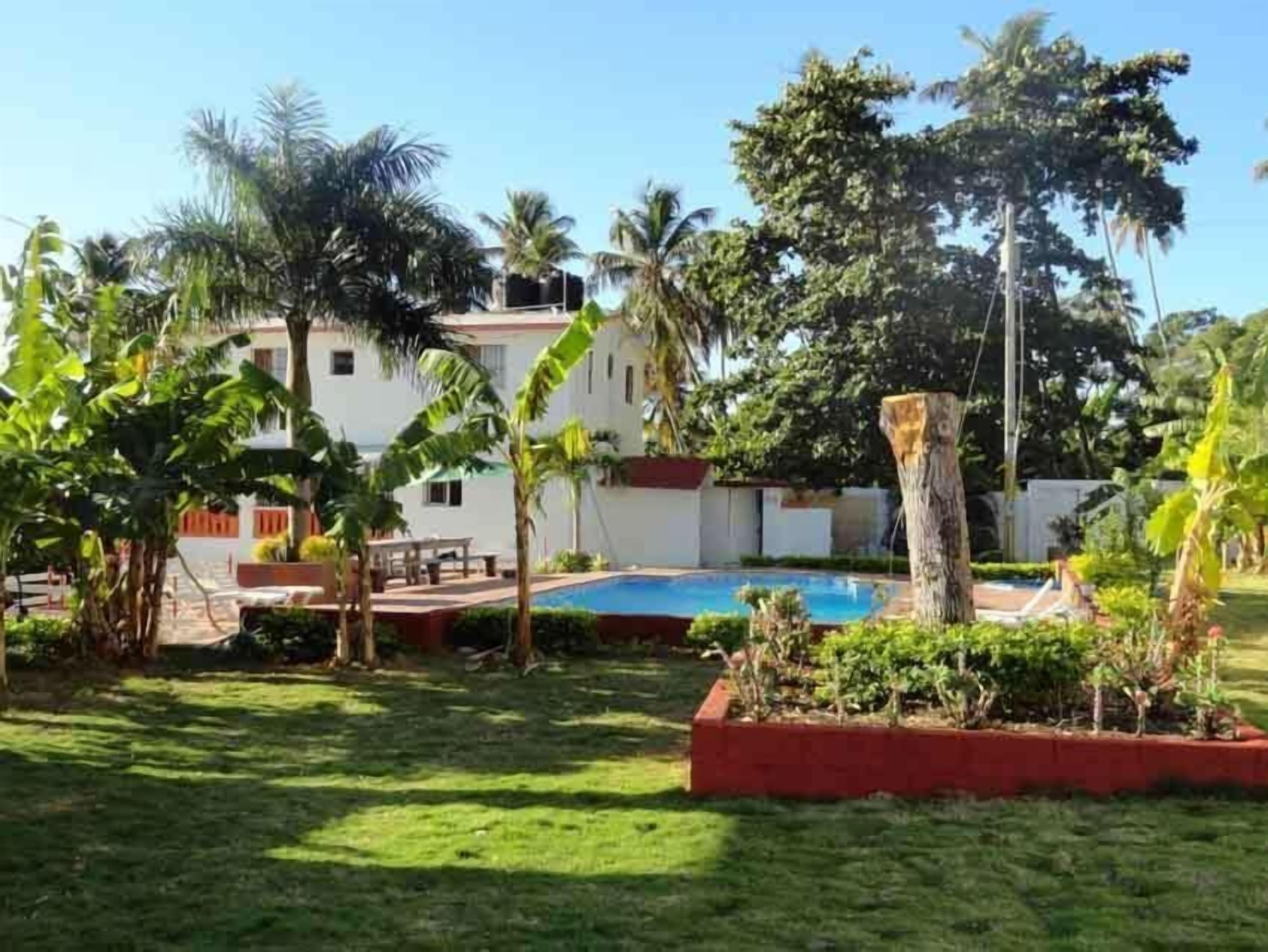 Una piscina rodeada de exuberante vegetación, un edificio blanco con detalles rojos y un césped bien cuidado.